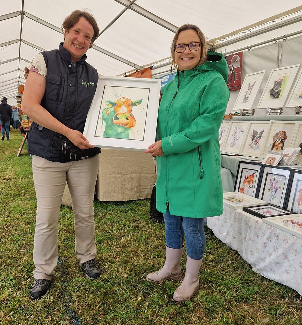 Two women holding framed animal painting at art fair.